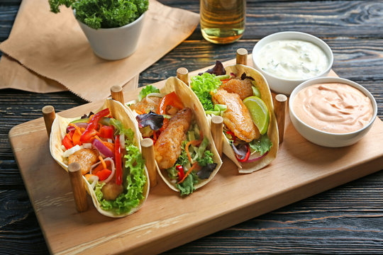 Wooden holder with delicious fish tacos and sauces on table