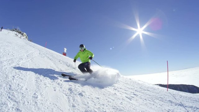 SLOW MOTION WIDE: Young skier stops, smiles and sprays snow to the camera on a piste at European ski resort in Alps. Perfect cold winter sunny day with a sun flare behind with no clouds