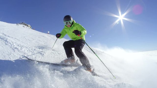SLOW MOTION WIDE: Young skier stops and smiles while sprays snow to the camera on a piste at European ski resort. Alps above the fog on a cloudless perfect winter day with sun flare behind