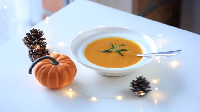 Pumpkin soup with rosemary in white plate near a pine cone with Fairy Ligths