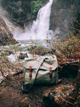 Old Swiss Army Bag With Leather Buckles On Ground In Front Of Flowing Waterfall