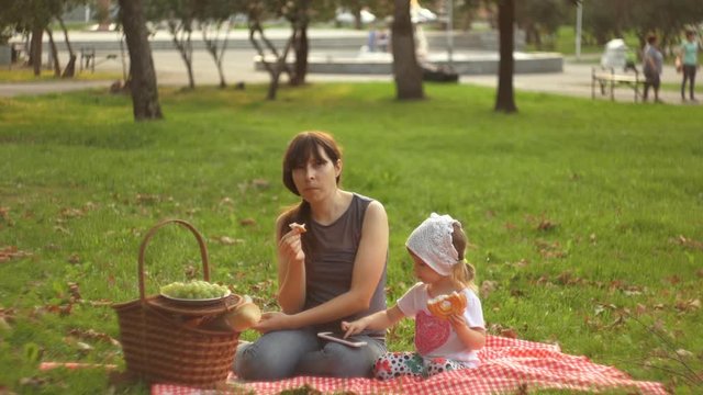 A family of two people, a mother and a small daughter, spends time together in a city park on a picnic. A young woman and a little girl are eating sweet burgers and enjoying a smartphone.