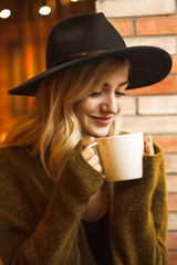 Beautiful young blonde woman sitting in the coffee bar in long autumn evening. She is wearing nice warm clothes and enjoying cup of coffee.