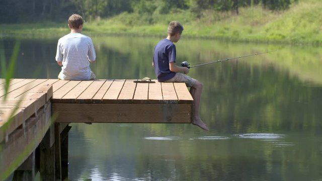 Two boys sitting on the end of a wooden jetty and fishing in a large pond surrounded by trees. Close view, 4K