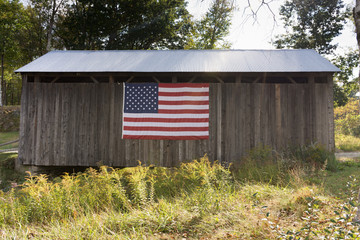 A weathered wood building and metal roof covering a bridge. The building is decorated with an American flag. Photographed in natural light. 