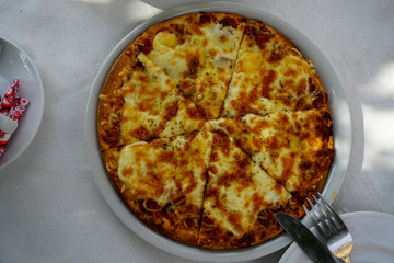 Fresh oven baked delicious Pizza Margherita top view showing mozzarella cheese serving on white round plate with fork and knife at street side cafe table on sunshine day