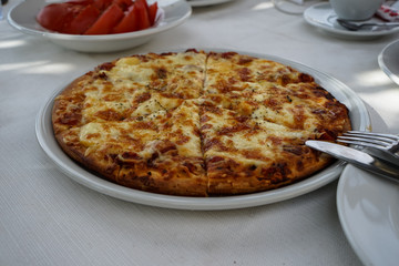Fresh oven baked delicious Pizza Margherita showing mozzarella cheese serving on white round plate with fork and knife at street side cafe table on sunshine day