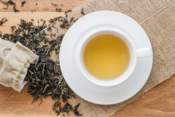 Cup of hot tea on sackcloth with dried tea leaves overflow form bag on wooden table with copy space from top view. Time to relax with hot tea in morning.