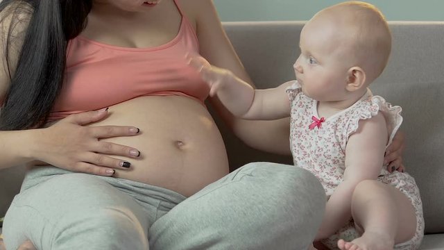 Baby girl and pregnant woman sitting on couch, child hitting stomach lightly