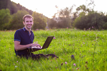 Young man sitting on green grass at sunset with laptop and working 