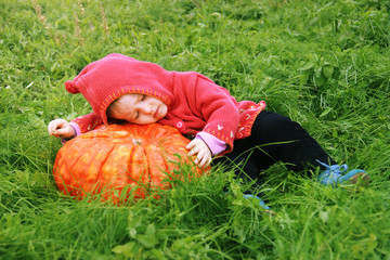 A small child in a red gnome costume falls asleep and sleeps on a huge pumpkin on the grass. Sunny warm day. copy space for text. The symbol and carnival costume of halloween