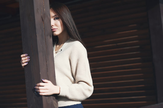 Portrait of a charming young woman in a knitted woolen beige sweater, on a wooden stall background. Hides half of face, and poses on camera