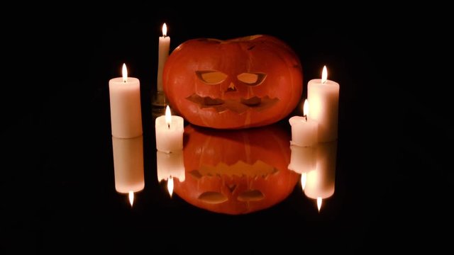 All Saints' Day,Halloween.Pumpkin with carved evil face and teeth shape saw lies on mirror surface,large and small candles burn around and inside pumpkin.Reflection pumpkin and candles in mirror.
