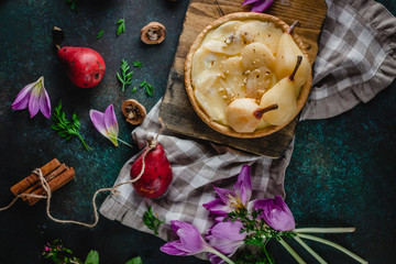 pear tart with nuts on wooden board with walnuts, fresh pears, rustic textile, cinnamon and purple flowers on green background