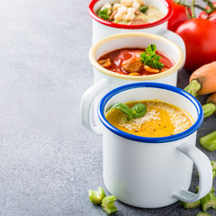 Assorted delicious homemade soups in enamel mugs with ingredients. Healthy food concept with copy space.