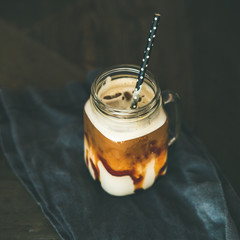 Iced caramel macciato coffee with milk in glass jar with straw over dark rustic wooden table background, selective focus, copy space, square crop. Cold sweet refreshing summer drink concept