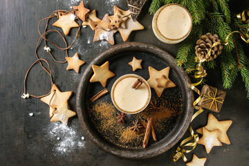 Eggnog Christmas milk cocktail with cinnamon, served in two glasses with shortbread star shape sugar cookies different size, decor toys, fir branch over dark texture background. Top view with space