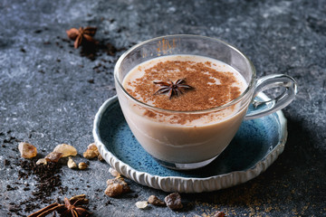 Glass cup of traditional indian masala chai tea on blue saucer with ingredients above. Cinnamon, cardamom, anise, sugar, black tea over dark texture background. Close up with space