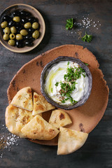 labneh middle eastern lebanese cream cheese dip with olive oil, salt, herbs served with olives, traditional pita bread on terracotta plate over dark texture metal background. Top view with space
