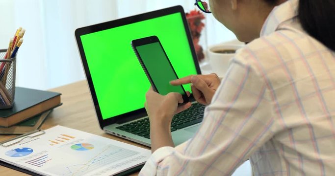 Asian woman hand holding cell phone.Phone and Laptop on desk with green screen.
