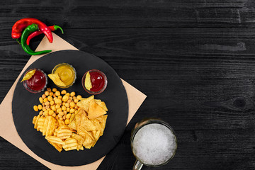 Overhead view of beer glass and snacks with sauce on black wooden table.