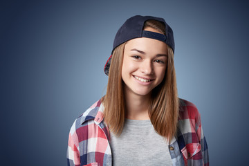 Closeup portrait of smiling teen girl wearing checkered shirt and baseball cap, over grey background