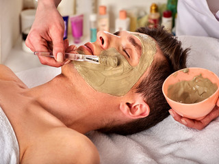 Mud facial mask of man in spa salon. Massage with clay full face. Girl on with therapy room....