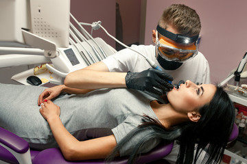 Young woman treats teeth in dental office. Male dentist treats her teeth with dental drill.