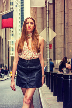 Young European Woman with long brown hair, traveling in New York, wearing light patterned, sleeveless deep v neck top, black leather skirt, walking on narrow vintage street of low Manhattan..