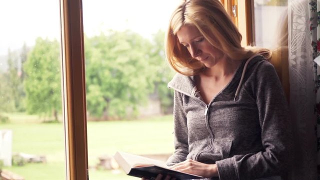Young, pretty woman reading book sitting by window at home
