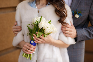 Winter wedding. Groom embracing bride in white fur coat holding bouquet of fresh winter or spring flowers. Winter love