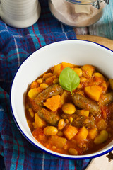 Butternut squash, pumpkin and bean stew with beef sausages. White bowl, dark blue tablecloth, wooden board.