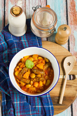 Butternut squash, pumpkin and bean stew with beef sausages. White bowl, dark blue tablecloth, wooden board.