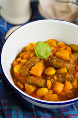 Butternut squash, pumpkin and bean stew with beef sausages. White bowl, dark blue tablecloth, wooden board.