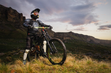 Naklejka na ściany i meble The rider in full protection on a mountain bike stands and looks at the sunset on the background of the rocks