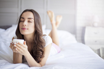 Attractive young smiling brunette woman lying in white bed and drinks a coffee in her bedroom.