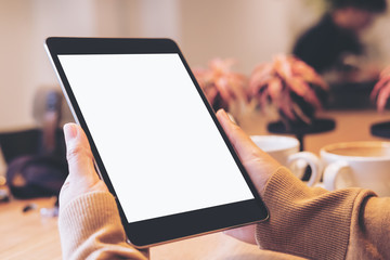 Mockup image of hands holding black tablet pc with blank white screen with coffee cups on wooden table in modern cafe