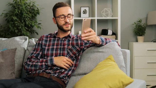 Handsome caucasian man in checked casula shirt and glasses is having a great time at home, sitting on the couch and videochatting with family at his free time. Indoor shot.