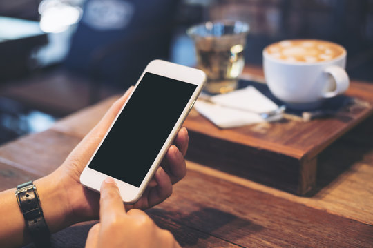 Mockup image of a woman's hands holding white mobile phone with blank black screen with coffee cup , tea and snack on wooden table in vintage cafe