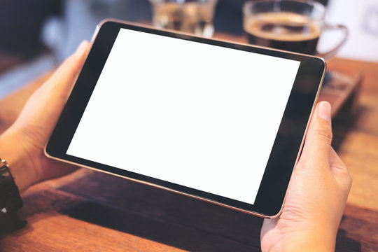 Mockup image of hands holding black tablet pc with blank white screen with coffee and tea on wooden table in cafe