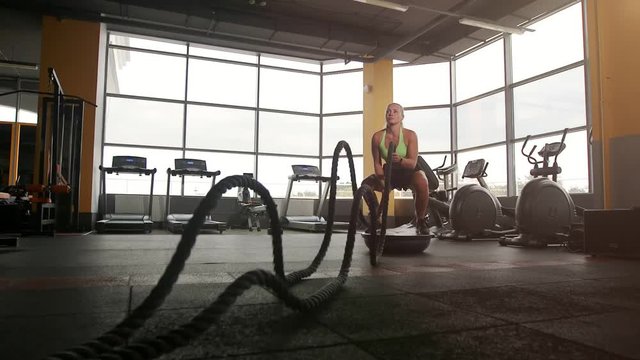 Athletic woman doing battle rope exercise in cross training gym during workout with personal coach