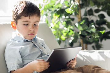 Little boy with digital tablet sitting on sofa, on home interior background