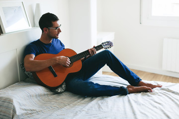 Young man playing guitar sitting on bed at home.