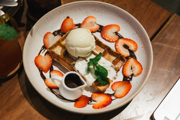 Top view of waffle topping with a scoop of vanilla ice cream, whip cream, sliced strawberry, mint leaves and chocolate sauce on white plate at dessert cafe in Bangkok, Thailand.