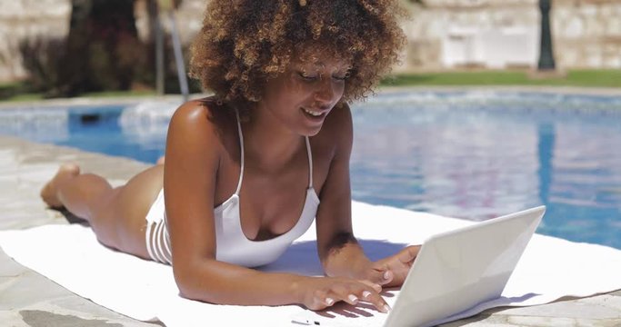 Young attractive woman lying on towel on poolside and typing laptop in sunny day.