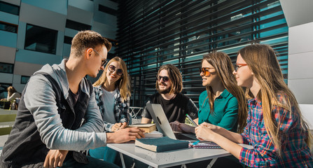 Casual stylish group of students have discussion on table with laptop and books before modern building, cafe sunny day