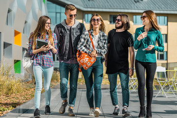 Fototapeta premium Casual style group of students walk along university modern building, have a pleasant talk, sunny day