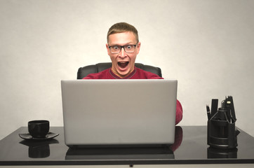 A young man in glasses and with wide open mouth working at a laptop at the table and in shock from what he saw. Man Looks amazing movie. Gamer playes in game. Special offer. Big sale.