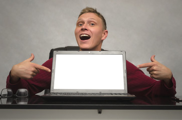 A smiling young man showing by fingers on blank screen of laptop computer on the table. Special offer. Big sale. Contact us. Advantages.