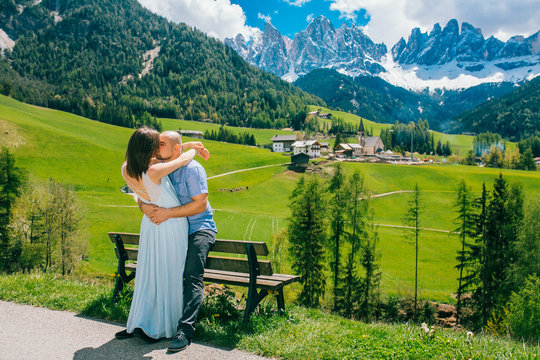 Young couple admires the view in relaxation. Famous best alpine place. Santa Maddalena village with magical Dolomites mountains in background, Val di Funes valley, Trentino Alto Adige Italy